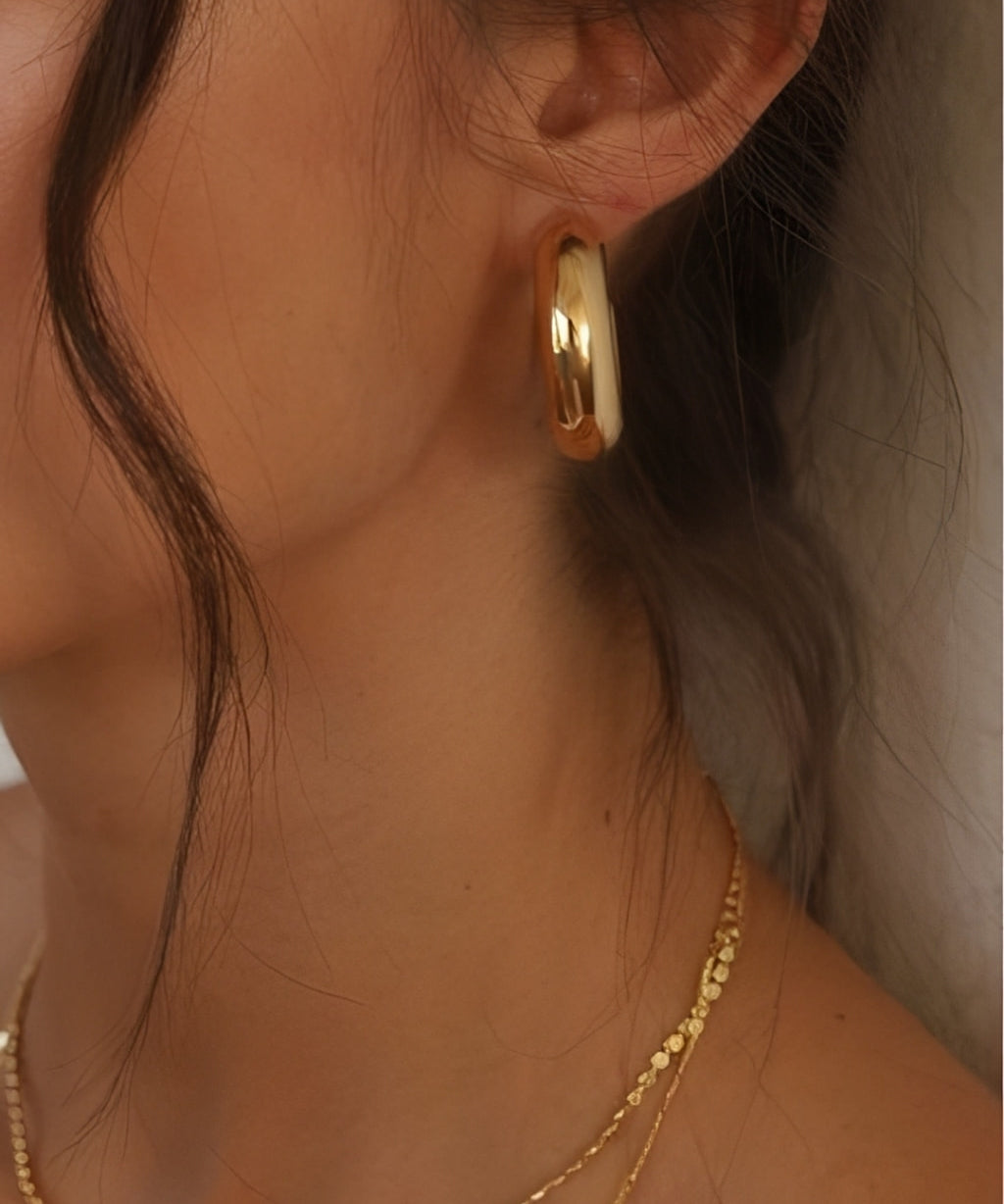 Ease Classic Oval Hoop Earrings in 14k gold plated finish worn on ear with smooth rounded oval silhouette.
