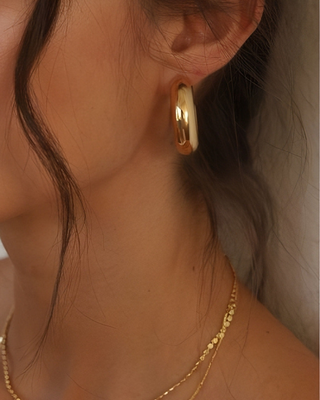 Ease Classic Oval Hoop Earrings in 14k gold plated finish worn on ear with smooth rounded oval silhouette.
