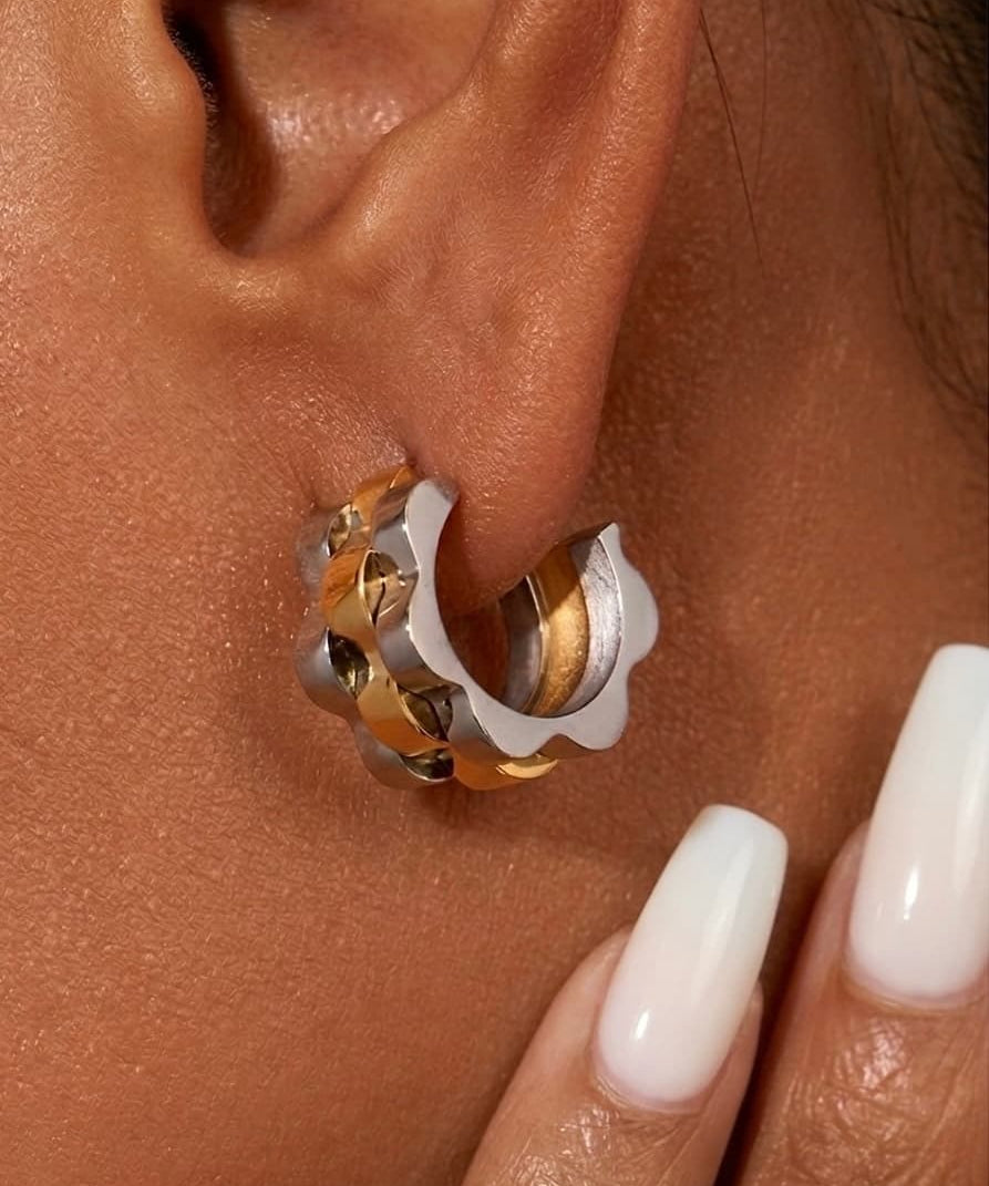 Close-up of wave cut hoop earrings in gold and silver worn on ear with milky white nails