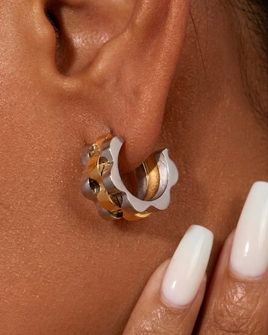 Close-up of wave cut hoop earrings in gold and silver worn on ear with milky white nails